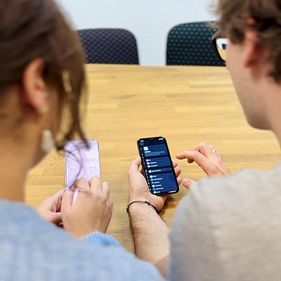 Zwei Personen halten jeweils ein iPhone in der Hand und bedienen die Einstellungen. Auf einem Gerät sind die iOS-Einstellungen im hellen Modus zu sehen, auf dem anderen im dunklen Modus.